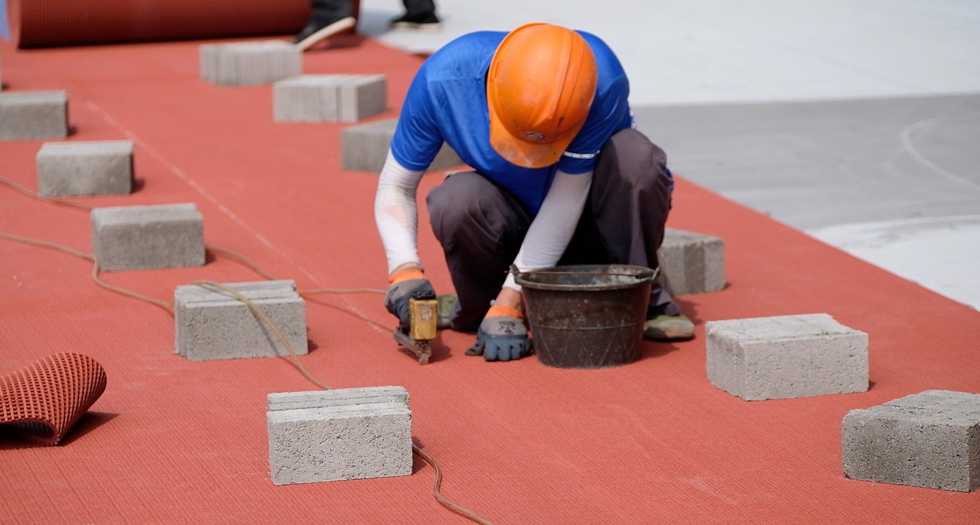 Installer working along a red rubber track sheet joint with temporary weights nearby